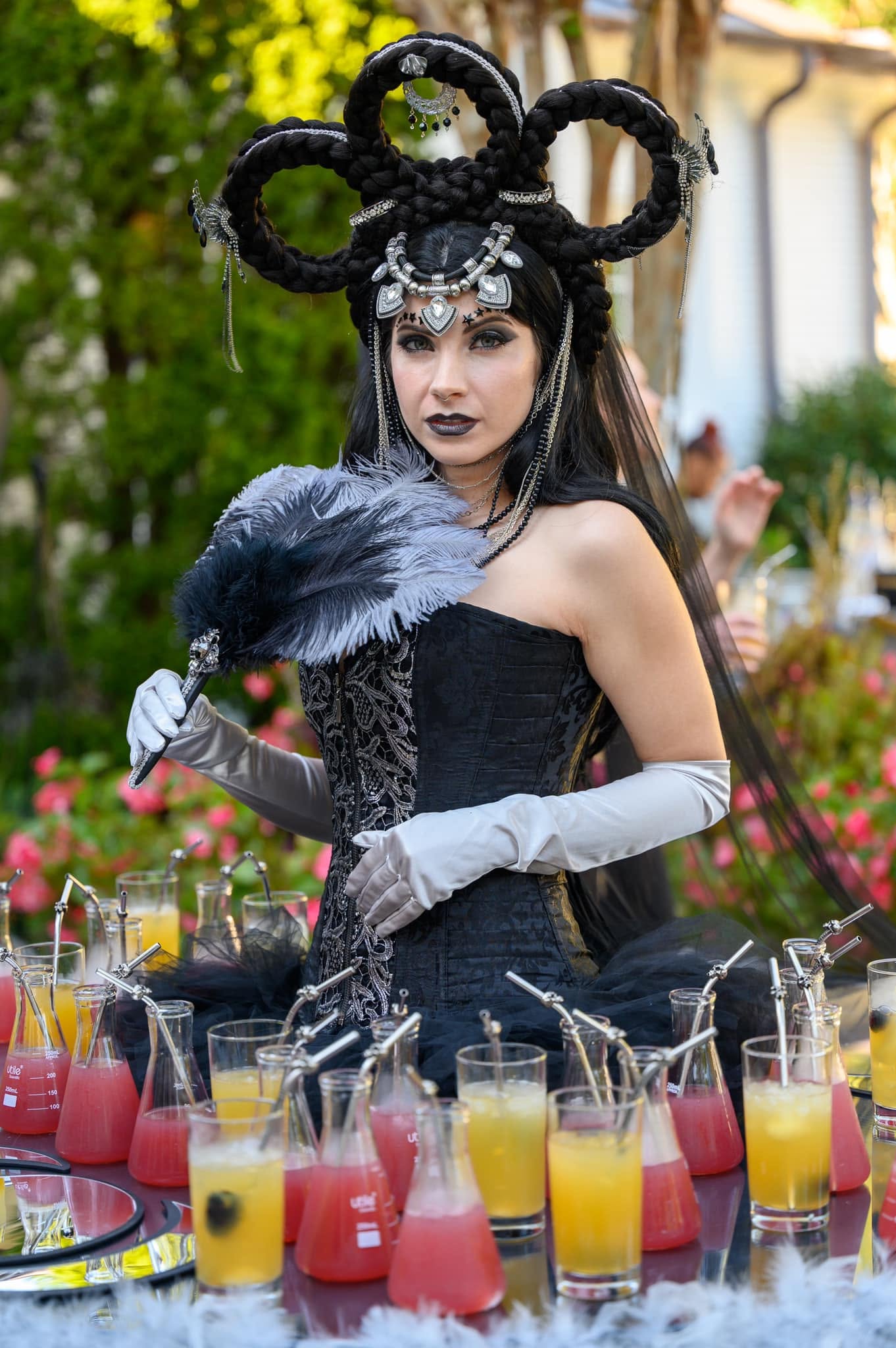 Character with a sculptural braided headpiece, feather fan, corset and gloves beside a beverage display