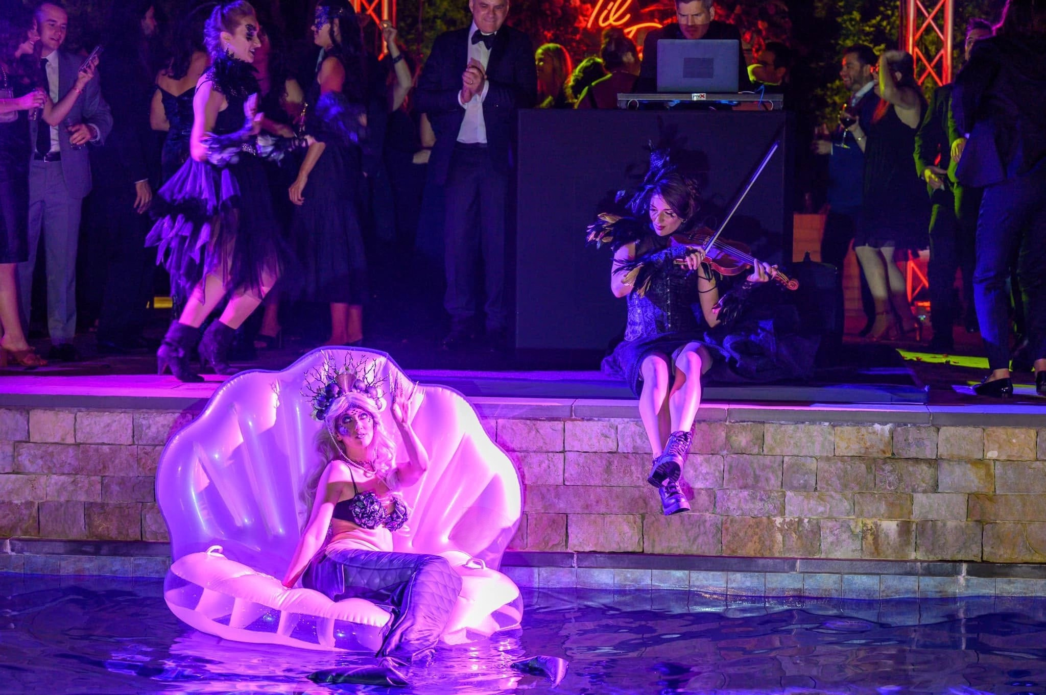 A mermaid in a shell float in the pool, a violinist at the pool edge, and costumed performers among guests at an illuminated Art To Life event